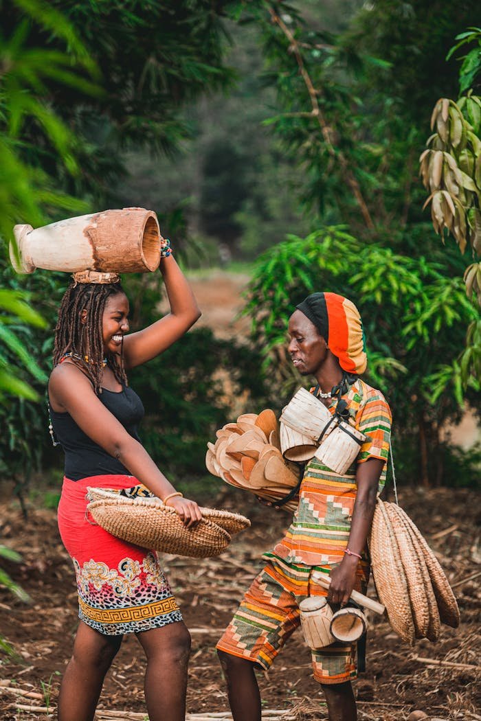 Two African women wearing colorful attire, carrying baskets and pots outdoors.