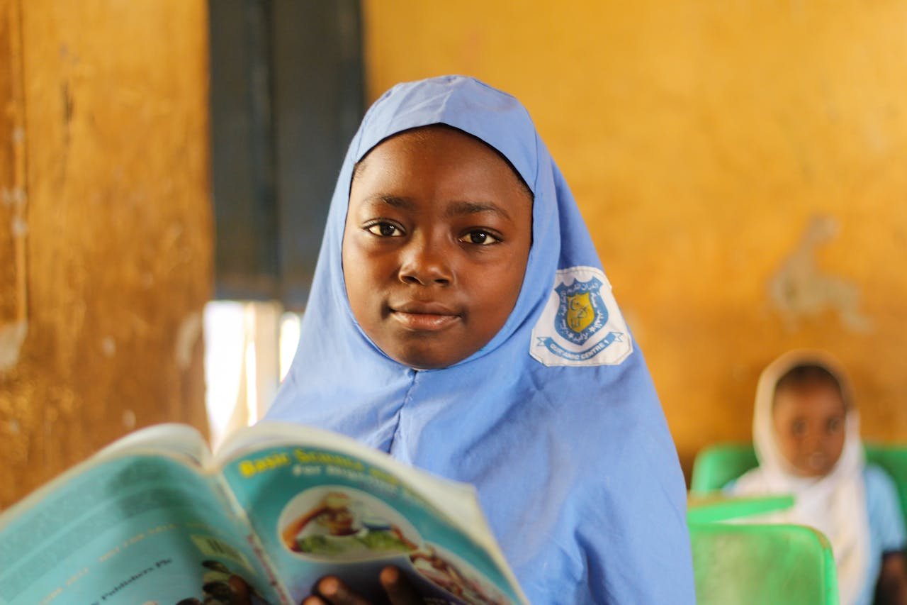 Young girl in blue hijab smiling while reading a book in a classroom setting.